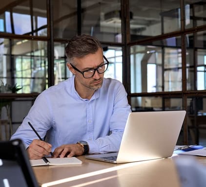 man writing notes in journal and looking at laptop